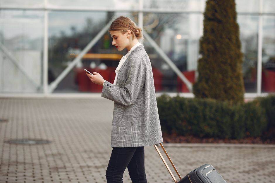 Stylish woman walking with suitcase while using smartphone outside a modern building.