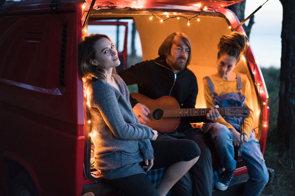 Friends enjoying a musical evening in a parked red van by the woods.