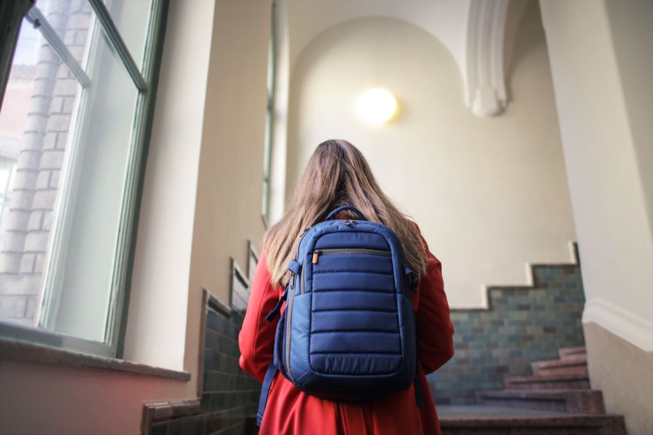 A woman in a red coat with a blue backpack climbs stairs inside a historic building.