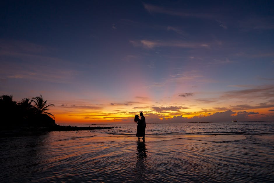 A romantic couple embraces on a beach at sunset, silhouetted against a colorful sky.