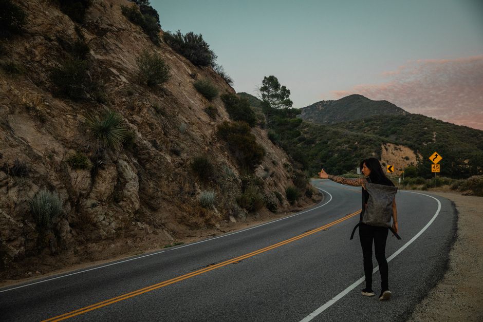 Woman with a backpack hitchhiking on a winding road at dusk.