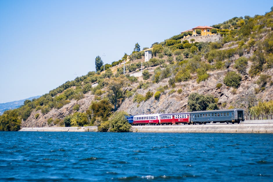 A colorful train travels alongside a picturesque hillside near a river, capturing nature's beauty and serene landscapes.