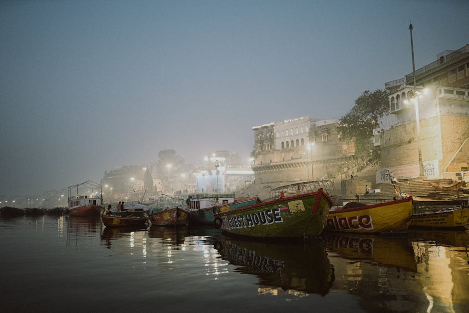 Misty evening view at Varanasi Ghats with colorful boats and illuminated buildings.